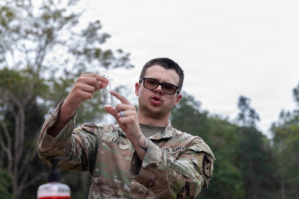 A U.S. Army soldier identified by the name tape 'LEE' is holding up and examining a small clear vial or container outdoors. The soldier is wearing Operational Camouflage Pattern (OCP) uniform and dark-framed sunglasses. The setting appears to be a wooded or semi-rural outdoor environment under overcast skies.