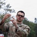 A U.S. Army soldier identified by the name tape 'LEE' is holding up and examining a small clear vial or container outdoors. The soldier is wearing Operational Camouflage Pattern (OCP) uniform and dark-framed sunglasses. The setting appears to be a wooded or semi-rural outdoor environment under overcast skies.
