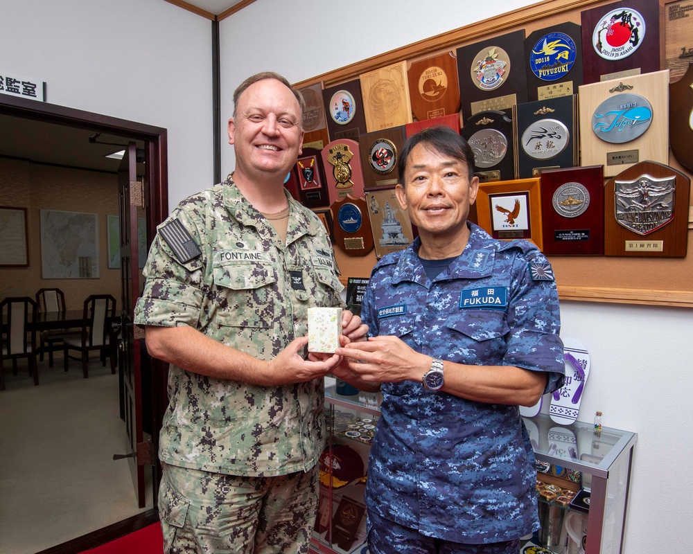Two military officers, one from the U.S. Navy and one from the Japan Maritime Self-Defense Force (JMSDF), are posing together while jointly holding a small decorative gift box. The setting appears to be an official military office decorated with numerous commemorative plaques, medallions, and unit insignia from both U.S. and Japanese naval forces. This appears to be a formal bilateral military exchange or courtesy visit.