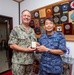 Two military officers, one from the U.S. Navy and one from the Japan Maritime Self-Defense Force (JMSDF), are posing together while jointly holding a small decorative gift box. The setting appears to be an official military office decorated with numerous commemorative plaques, medallions, and unit insignia from both U.S. and Japanese naval forces. This appears to be a formal bilateral military exchange or courtesy visit.