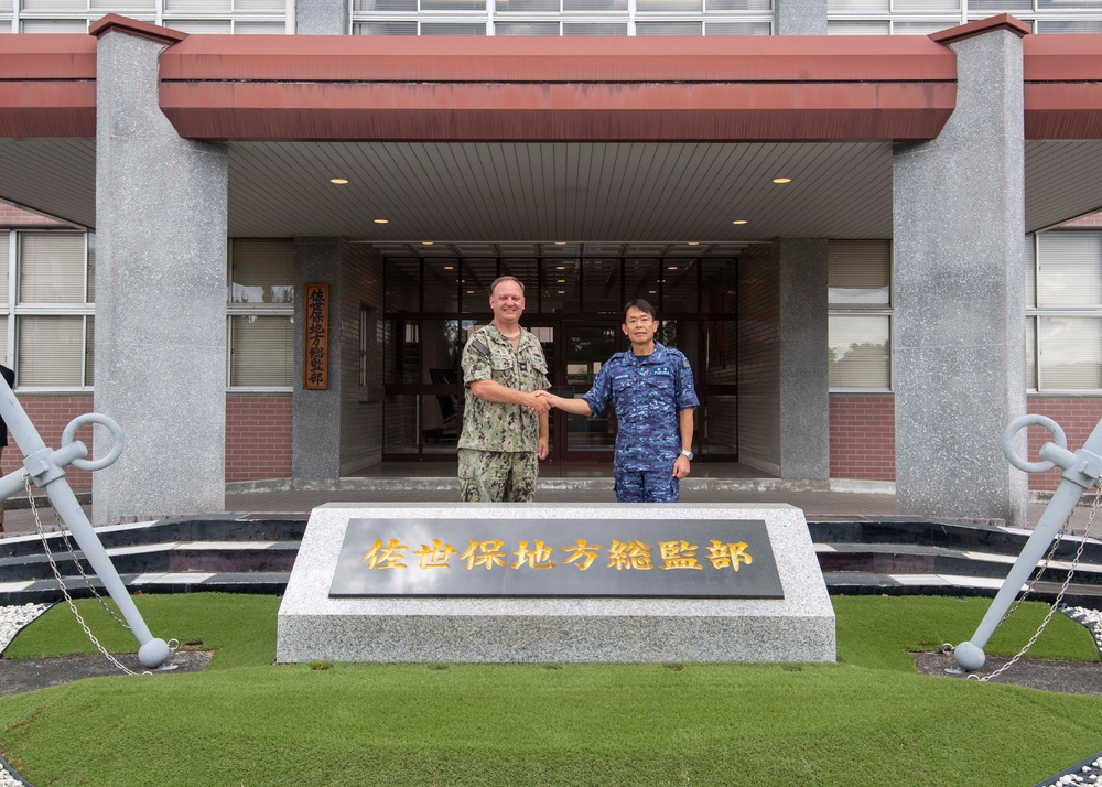 Two military officers, one in U.S. Navy digital camouflage (NWU) and one in Japan Maritime Self-Defense Force (JMSDF) blue camouflage uniform, are shaking hands in front of a building entrance. The stone monument in the foreground displays Japanese characters reading '佐世保地方総監部' (Sasebo District Headquarters). The setting appears to be the JMSDF Sasebo District Headquarters in Sasebo, Japan, suggesting a formal bilateral military meeting.