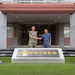 Two military officers, one in U.S. Navy digital camouflage (NWU) and one in Japan Maritime Self-Defense Force (JMSDF) blue camouflage uniform, are shaking hands in front of a building entrance. The stone monument in the foreground displays Japanese characters reading '佐世保地方総監部' (Sasebo District Headquarters). The setting appears to be the JMSDF Sasebo District Headquarters in Sasebo, Japan, suggesting a formal bilateral military meeting.