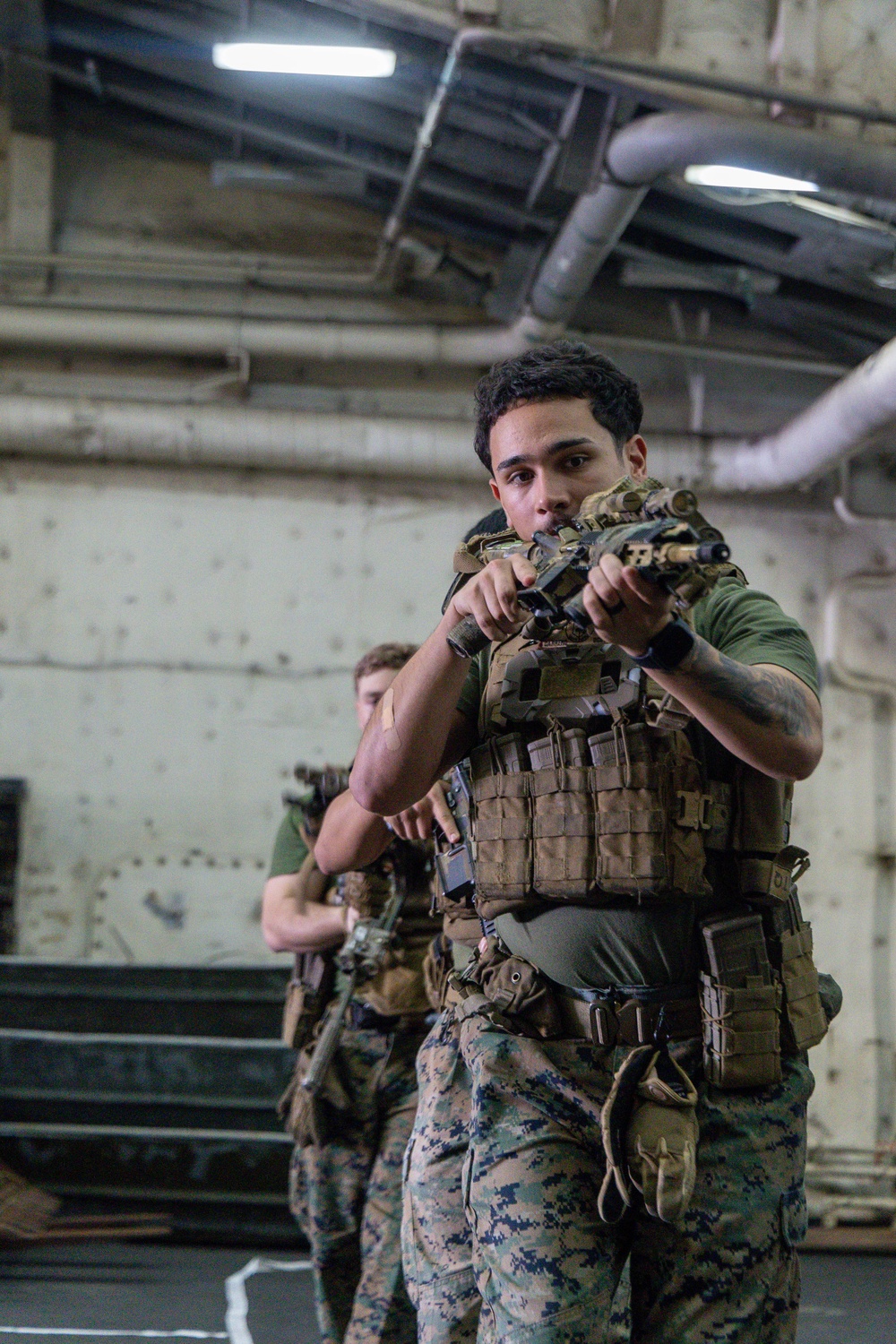 Two military personnel, appearing to be U.S. Marines based on MARPAT digital camouflage uniforms, are conducting indoor close-quarters battle (CQB) training in what appears to be the interior of a naval vessel. The foreground Marine is in a low-ready or aimed firing stance with a rifle equipped with an optic, wearing a plate carrier with MOLLE pouches. The environment features exposed pipes, riveted metal walls, and fluorescent lighting consistent with a ship's well deck or hangar bay.
