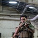 Two military personnel, appearing to be U.S. Marines based on MARPAT digital camouflage uniforms, are conducting indoor close-quarters battle (CQB) training in what appears to be the interior of a naval vessel. The foreground Marine is in a low-ready or aimed firing stance with a rifle equipped with an optic, wearing a plate carrier with MOLLE pouches. The environment features exposed pipes, riveted metal walls, and fluorescent lighting consistent with a ship's well deck or hangar bay.