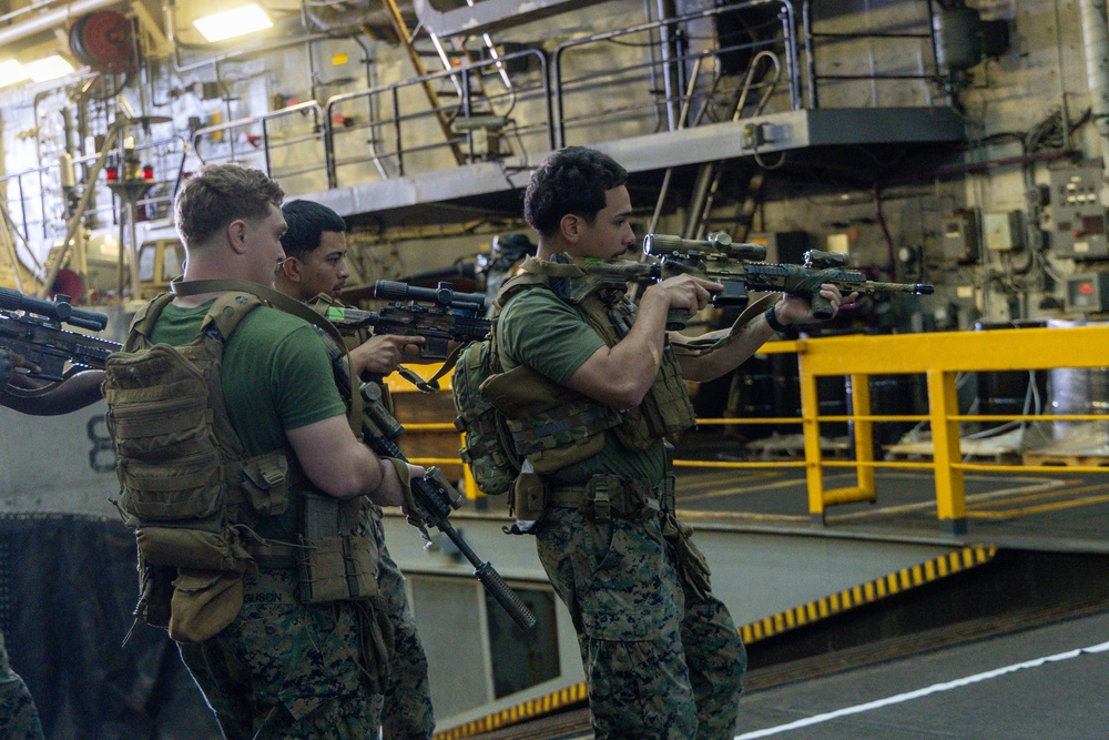 Three military personnel in camouflage uniforms and tactical gear are conducting what appears to be a training exercise aboard a naval vessel. They are positioned in a tactical formation, aiming rifles equipped with scopes and accessories. The setting is an interior deck or well deck of a large military ship.