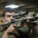 A young male in military/tactical gear is holding up a heavily accessorized assault rifle in what appears to be an indoor setting. The rifle is fitted with multiple optics, a laser aiming device, and other tactical attachments in a tan/camouflage finish. The subject is wearing a green t-shirt and a tactical vest.