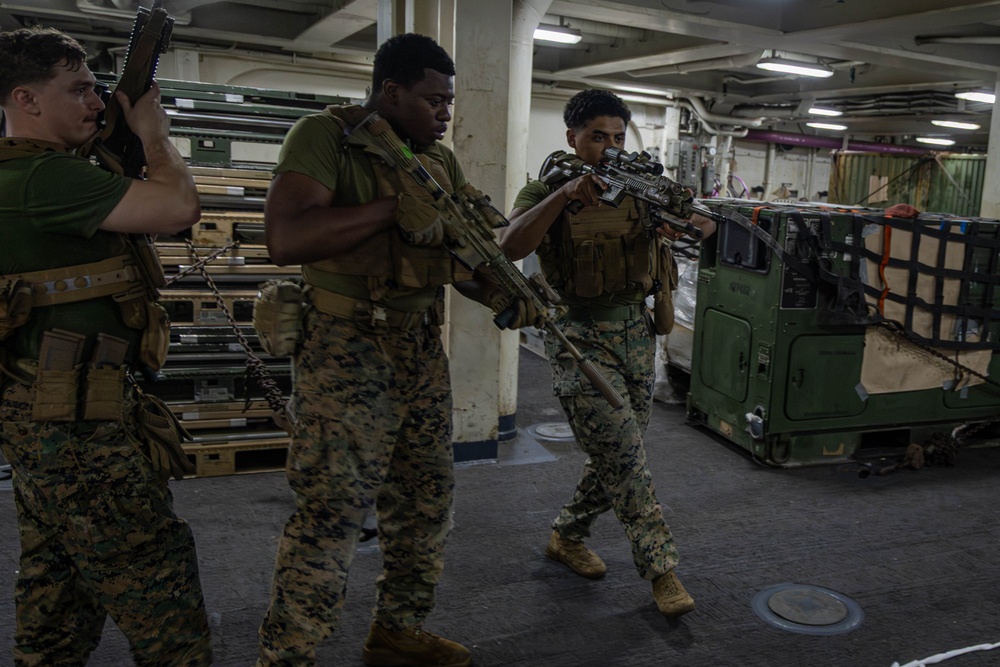 Three military personnel in MARPAT camouflage uniforms and tactical gear are conducting what appears to be a clearing or room-entry drill inside the cargo hold of a naval vessel. They are armed with rifles and wearing plate carrier vests, moving in a tactical formation. The setting shows a large interior ship deck with military vehicles, pallets, and equipment secured in the background.