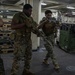 Three military personnel in MARPAT camouflage uniforms and tactical gear are conducting what appears to be a clearing or room-entry drill inside the cargo hold of a naval vessel. They are armed with rifles and wearing plate carrier vests, moving in a tactical formation. The setting shows a large interior ship deck with military vehicles, pallets, and equipment secured in the background.