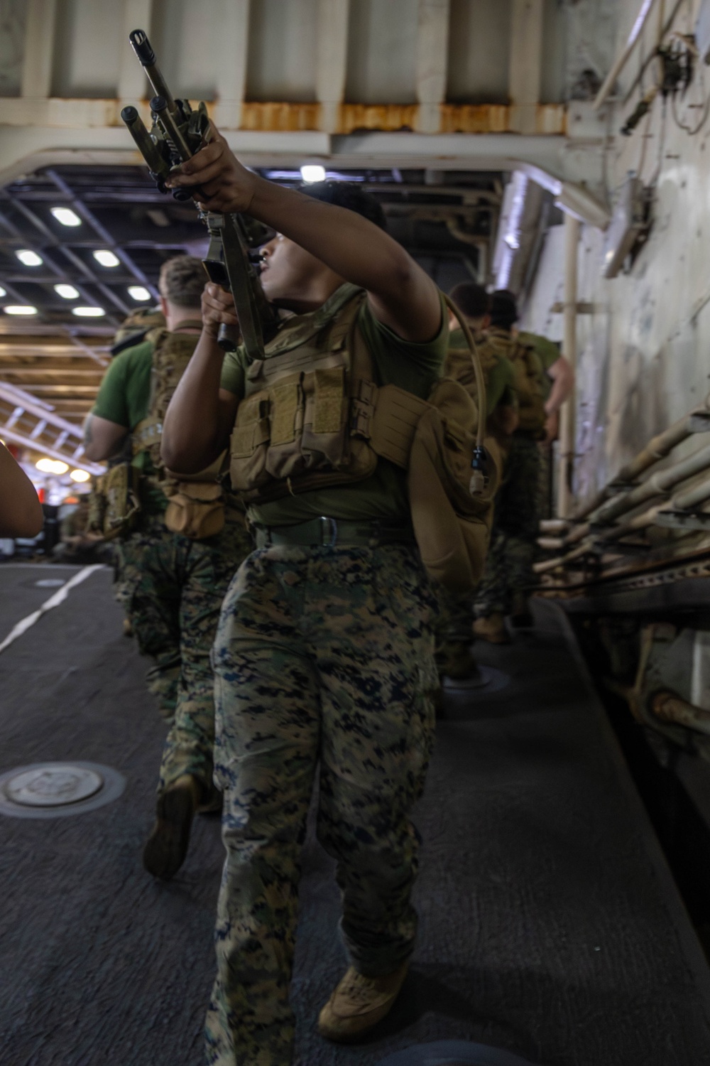 A group of military personnel in MARPAT camouflage uniforms and tactical vests are moving through what appears to be the interior deck of a naval vessel, likely a well deck or vehicle bay. The foreground figure is performing a barrel/chamber inspection by looking down the muzzle of a rifle held upward. The setting and uniforms are consistent with U.S. Marine Corps operations aboard an amphibious assault ship.