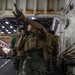 A group of military personnel in MARPAT camouflage uniforms and tactical vests are moving through what appears to be the interior deck of a naval vessel, likely a well deck or vehicle bay. The foreground figure is performing a barrel/chamber inspection by looking down the muzzle of a rifle held upward. The setting and uniforms are consistent with U.S. Marine Corps operations aboard an amphibious assault ship.