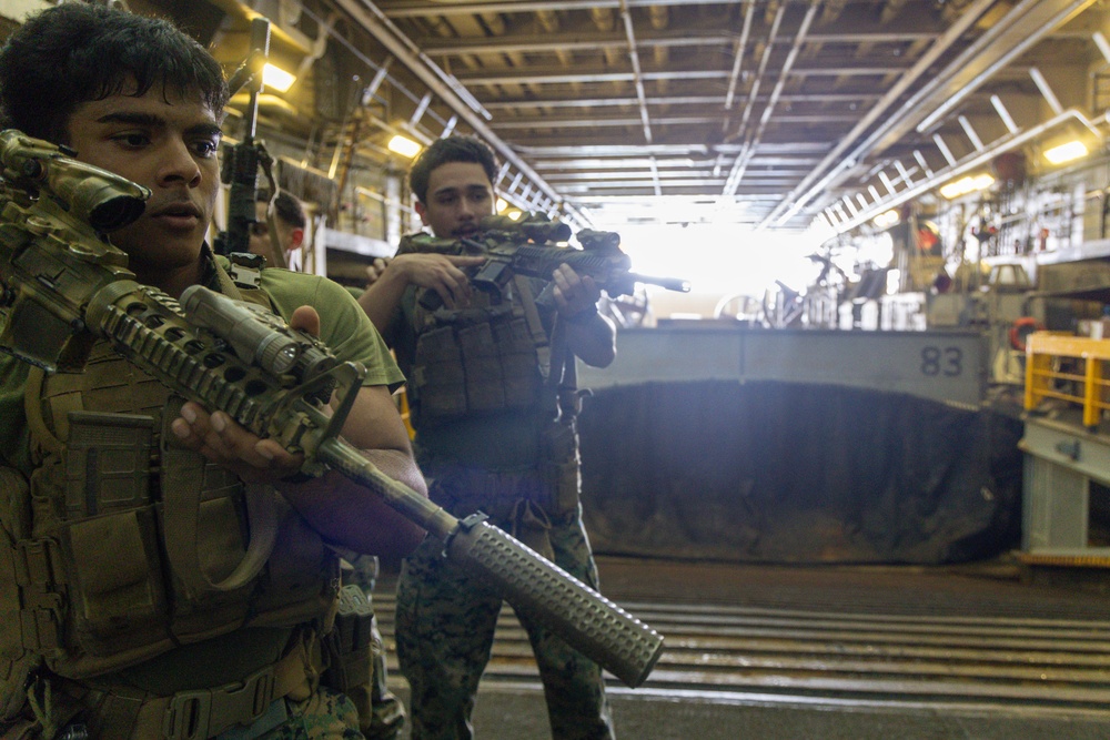 Two armed military personnel in tactical gear are positioned inside what appears to be a naval vessel's well deck, holding rifles in a ready stance. Behind them, a tracked amphibious assault vehicle marked with the number '83' is visible. The setting suggests a training exercise or operational readiness drill aboard a ship.