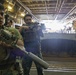 Two armed military personnel in tactical gear are positioned inside what appears to be a naval vessel's well deck, holding rifles in a ready stance. Behind them, a tracked amphibious assault vehicle marked with the number '83' is visible. The setting suggests a training exercise or operational readiness drill aboard a ship.