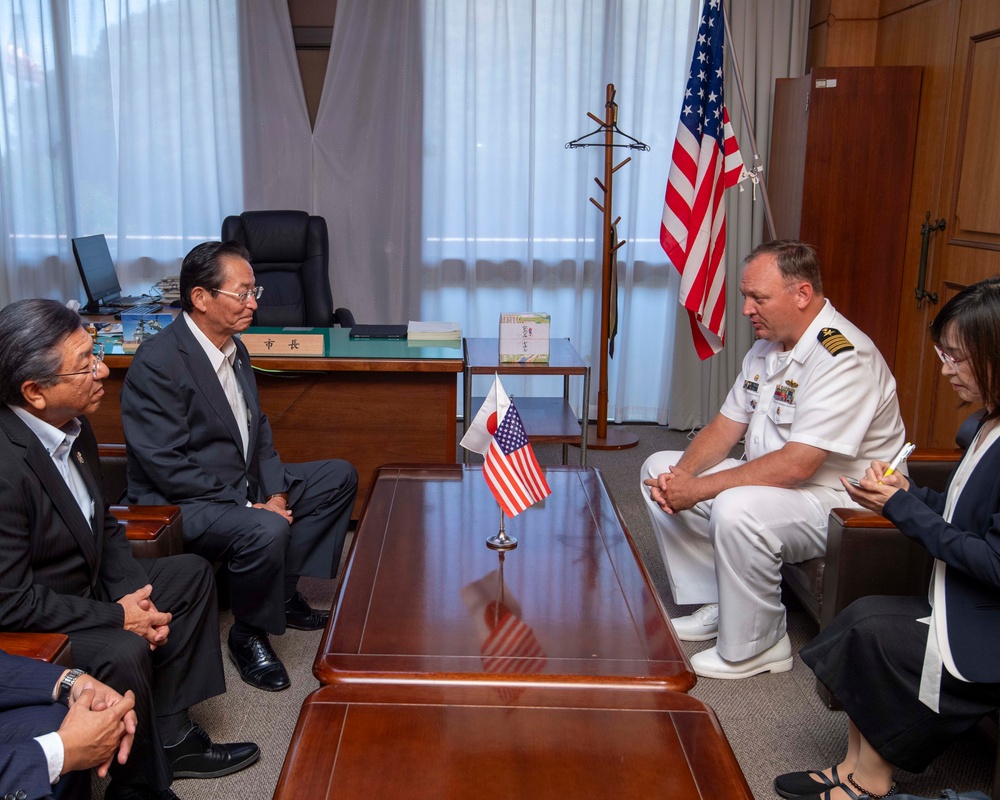 A formal diplomatic meeting is taking place between a U.S. Navy officer in white dress uniform and Japanese officials in a formal office setting. Small desktop flags of both the United States and Japan are placed on the conference table, and a full-sized American flag stands in the background. A woman on the right appears to be taking notes, suggesting this is an official bilateral meeting.