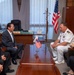 A formal diplomatic meeting is taking place between a U.S. Navy officer in white dress uniform and Japanese officials in a formal office setting. Small desktop flags of both the United States and Japan are placed on the conference table, and a full-sized American flag stands in the background. A woman on the right appears to be taking notes, suggesting this is an official bilateral meeting.