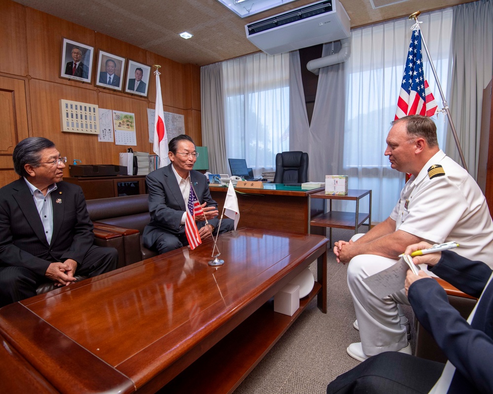 A formal bilateral meeting is taking place in a Japanese official's office, with two Japanese civilians and a U.S. Navy officer seated around a wooden conference table. Small U.S. and Japanese desk flags are displayed on the table, with full-sized versions of both flags visible in the background. A fourth person, partially visible at the edge of the frame, appears to be taking notes.