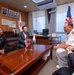 A formal bilateral meeting is taking place in a Japanese official's office, with two Japanese civilians and a U.S. Navy officer seated around a wooden conference table. Small U.S. and Japanese desk flags are displayed on the table, with full-sized versions of both flags visible in the background. A fourth person, partially visible at the edge of the frame, appears to be taking notes.