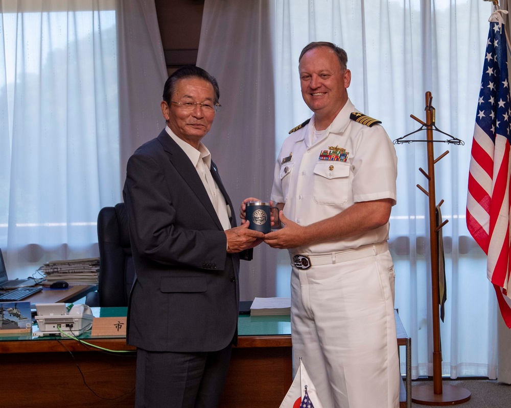 A US Navy officer in white dress uniform is presenting or exchanging a commemorative gift with a civilian man in a dark suit, likely in an official office setting. The presence of an American flag, a small Japanese flag, and what appears to be an official desk nameplate suggests this is a formal diplomatic or courtesy visit. The exchange appears to be a token of goodwill between military and local civilian leadership.