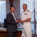 A US Navy officer in white dress uniform is presenting or exchanging a commemorative gift with a civilian man in a dark suit, likely in an official office setting. The presence of an American flag, a small Japanese flag, and what appears to be an official desk nameplate suggests this is a formal diplomatic or courtesy visit. The exchange appears to be a token of goodwill between military and local civilian leadership.
