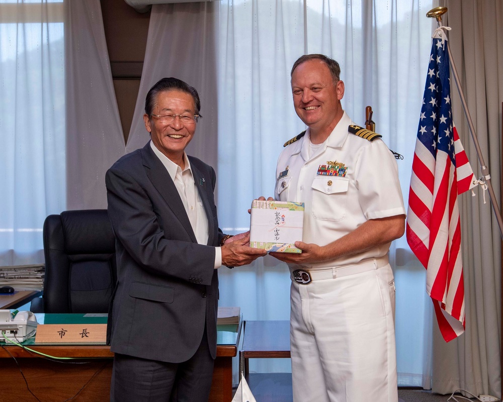 A formal meeting between a Japanese civilian official and a U.S. Navy officer, likely a Captain based on the four gold stripes on his shoulder boards. The two men are jointly holding a wrapped gift or ceremonial item, suggesting an official exchange or presentation ceremony. A nameplate reading '市長' (Mayor) is visible on the desk behind them, indicating the civilian is a city mayor.