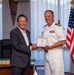 A formal meeting between a Japanese civilian official and a U.S. Navy officer, likely a Captain based on the four gold stripes on his shoulder boards. The two men are jointly holding a wrapped gift or ceremonial item, suggesting an official exchange or presentation ceremony. A nameplate reading '市長' (Mayor) is visible on the desk behind them, indicating the civilian is a city mayor.