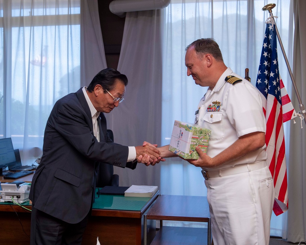A formal exchange ceremony between a civilian official in a dark suit and a U.S. Navy officer in white dress uniform. The naval officer is receiving or exchanging a wrapped gift/package with Japanese text visible on it. The setting appears to be an official office environment with an American flag displayed prominently in the background.