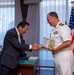 A formal exchange ceremony between a civilian official in a dark suit and a U.S. Navy officer in white dress uniform. The naval officer is receiving or exchanging a wrapped gift/package with Japanese text visible on it. The setting appears to be an official office environment with an American flag displayed prominently in the background.