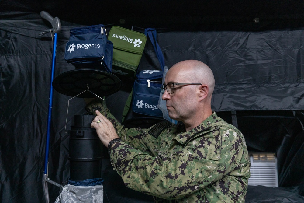 A bald male in military camouflage uniform (appears to be US Navy NWU Type III digital camo) is operating or adjusting a Biogents mosquito trap device inside what appears to be a field tent or temporary shelter. Multiple Biogents-branded carrying cases in navy blue, olive green, and black are visible hanging in the background. The setting suggests a military entomology or vector control operation.