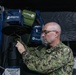 A bald male in military camouflage uniform (appears to be US Navy NWU Type III digital camo) is operating or adjusting a Biogents mosquito trap device inside what appears to be a field tent or temporary shelter. Multiple Biogents-branded carrying cases in navy blue, olive green, and black are visible hanging in the background. The setting suggests a military entomology or vector control operation.