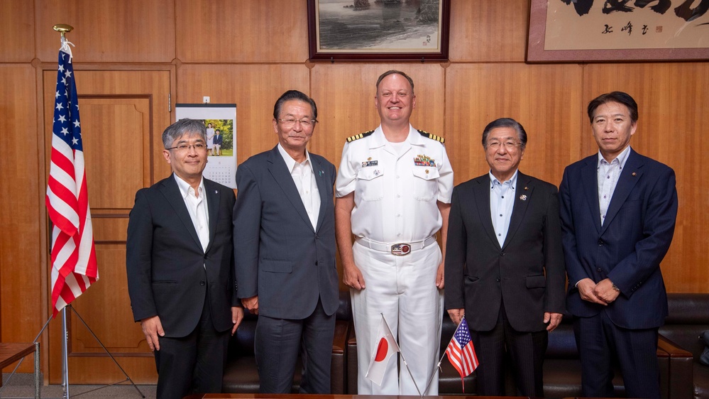 A formal group photograph featuring five individuals posing together in what appears to be an official office setting. One individual is wearing a U.S. Navy officer's white dress uniform, while the other four are dressed in civilian business attire. Both a large U.S. flag and small Japanese and American desk flags are visible, suggesting a bilateral diplomatic or military meeting.
