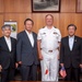 A formal group photograph featuring five individuals posing together in what appears to be an official office setting. One individual is wearing a U.S. Navy officer's white dress uniform, while the other four are dressed in civilian business attire. Both a large U.S. flag and small Japanese and American desk flags are visible, suggesting a bilateral diplomatic or military meeting.
