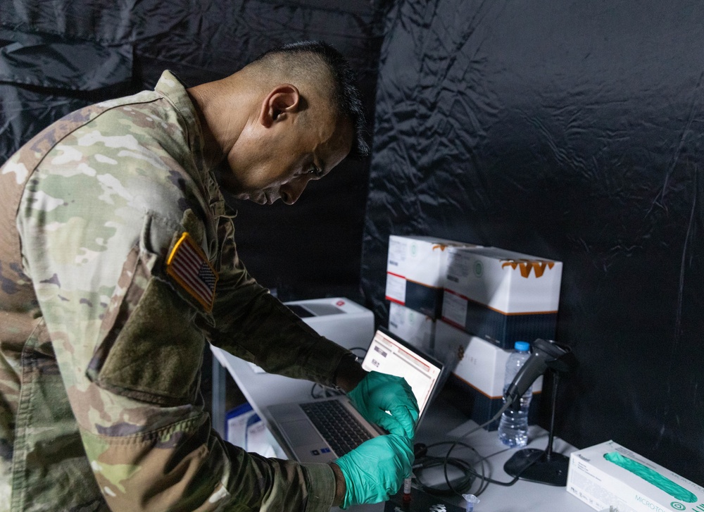 A U.S. Army soldier wearing multicam/OCP camouflage uniform with an American flag patch is working at a field processing station, scanning or reviewing a document/label while wearing teal nitrile gloves. The setting appears to be a temporary field tent or enclosure with dark walls, equipped with a laptop, barcode scanner, medical supply boxes, and specimen tubes. This appears to be a military medical or testing operation, likely a COVID-19 testing or vaccination site.