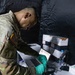 A U.S. Army soldier wearing multicam/OCP camouflage uniform with an American flag patch is working at a field processing station, scanning or reviewing a document/label while wearing teal nitrile gloves. The setting appears to be a temporary field tent or enclosure with dark walls, equipped with a laptop, barcode scanner, medical supply boxes, and specimen tubes. This appears to be a military medical or testing operation, likely a COVID-19 testing or vaccination site.