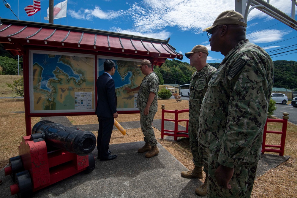 Three U.S. Navy personnel in digital camouflage uniforms are meeting with a civilian in a business suit at an outdoor map display board with Japanese architectural styling. The group appears to be reviewing a nautical or regional map, likely at a U.S. naval installation in Japan, as indicated by both the American and Japanese flags visible in the background. A historical black cannon on a red mount is displayed in the foreground as a decorative or commemorative artifact.