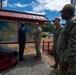 Three U.S. Navy personnel in digital camouflage uniforms are meeting with a civilian in a business suit at an outdoor map display board with Japanese architectural styling. The group appears to be reviewing a nautical or regional map, likely at a U.S. naval installation in Japan, as indicated by both the American and Japanese flags visible in the background. A historical black cannon on a red mount is displayed in the foreground as a decorative or commemorative artifact.