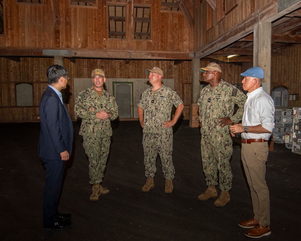 Five individuals are gathered inside a large wooden structure, appearing to be in a briefing or discussion. Three are wearing U.S. Navy camouflage uniforms (NWU Type III), one is in civilian business attire (dark suit), and one is in civilian casual attire (white shirt, khaki pants) with a light blue cap. The setting appears to be a historic or older wooden warehouse or barn-like facility.