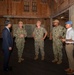 Five individuals are gathered inside a large wooden structure, appearing to be in a briefing or discussion. Three are wearing U.S. Navy camouflage uniforms (NWU Type III), one is in civilian business attire (dark suit), and one is in civilian casual attire (white shirt, khaki pants) with a light blue cap. The setting appears to be a historic or older wooden warehouse or barn-like facility.