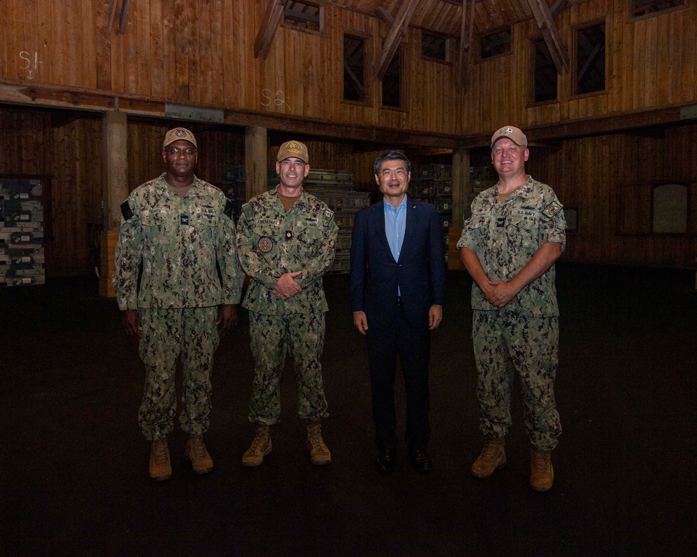 Four individuals pose for a formal group photograph inside a large wooden structure. Three of the individuals are wearing U.S. Navy NWU Type III (woodland camouflage) uniforms with tan boots and caps, while one civilian in a blue suit stands among them. The setting appears to be a military facility or warehouse with wooden walls and stored equipment visible in the background.