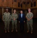 Four individuals pose for a formal group photograph inside a large wooden structure. Three of the individuals are wearing U.S. Navy NWU Type III (woodland camouflage) uniforms with tan boots and caps, while one civilian in a blue suit stands among them. The setting appears to be a military facility or warehouse with wooden walls and stored equipment visible in the background.