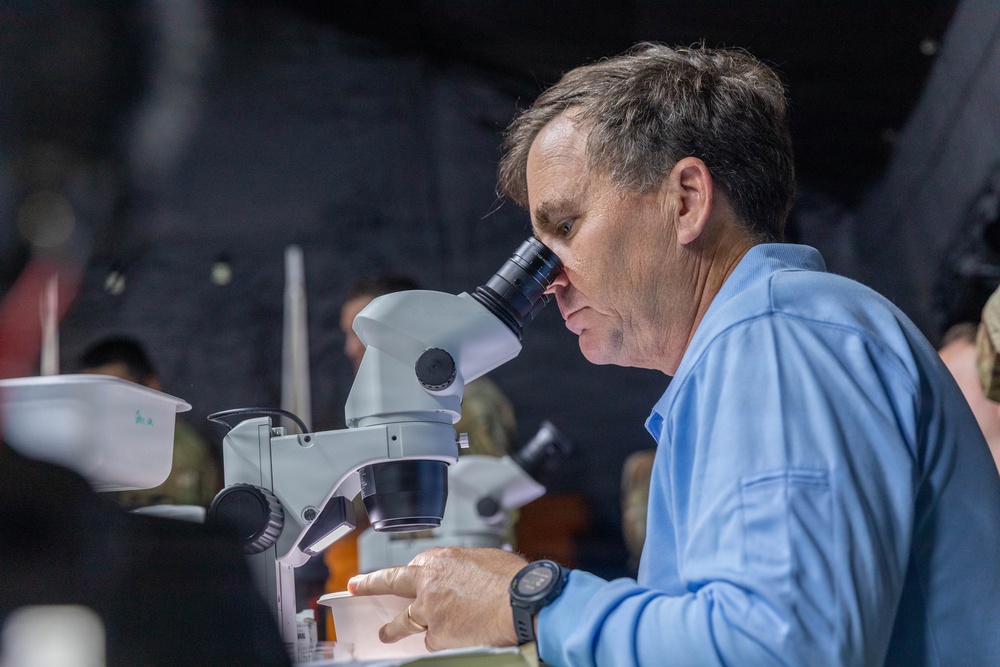 A middle-aged man in a light blue long-sleeve shirt is looking through a stereo/dissecting microscope. He appears to be in a semi-dark environment, possibly a field lab or demonstration setting, with other people and equipment visible in the background. His focused posture and the professional equipment suggest a scientific examination or analysis activity.