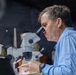 A middle-aged man in a light blue long-sleeve shirt is looking through a stereo/dissecting microscope. He appears to be in a semi-dark environment, possibly a field lab or demonstration setting, with other people and equipment visible in the background. His focused posture and the professional equipment suggest a scientific examination or analysis activity.