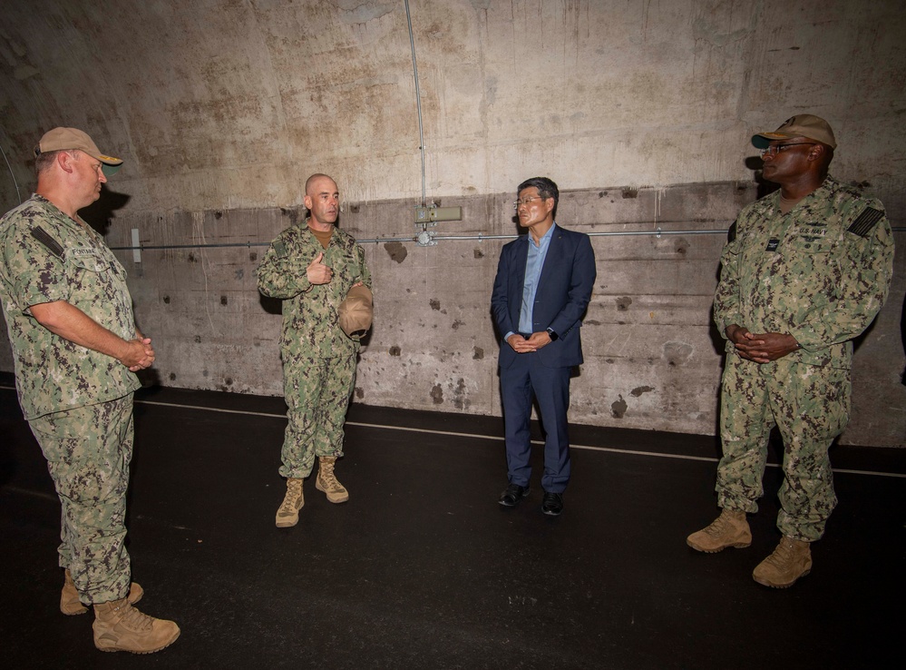 Four individuals are gathered in what appears to be a large concrete bunker or industrial facility. Three are wearing U.S. Navy camouflage (NWU Type III) uniforms, while one civilian in a navy blue suit is present, suggesting an official visit or briefing. The group appears to be engaged in a discussion, with one uniformed individual gesturing while speaking.