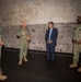 Four individuals are gathered in what appears to be a large concrete bunker or industrial facility. Three are wearing U.S. Navy camouflage (NWU Type III) uniforms, while one civilian in a navy blue suit is present, suggesting an official visit or briefing. The group appears to be engaged in a discussion, with one uniformed individual gesturing while speaking.