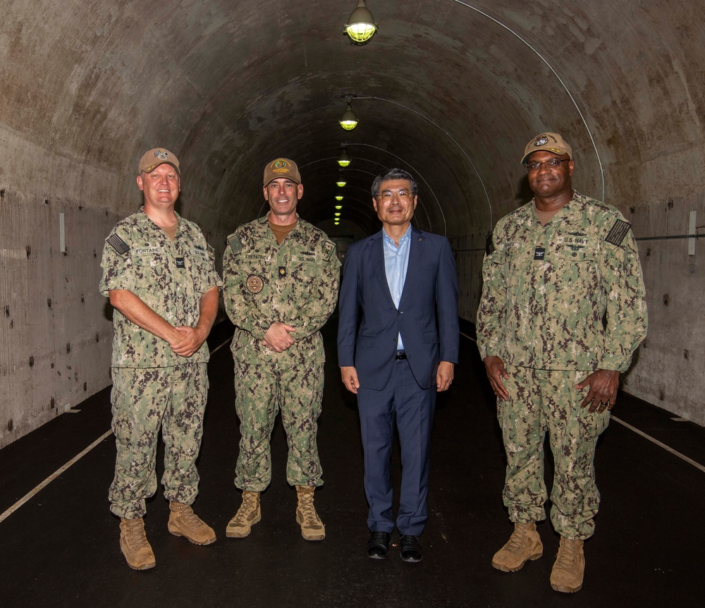 Four individuals pose for a formal photograph inside a large concrete tunnel. Three are wearing U.S. Navy NWU Type III (woodland camouflage) uniforms, while one civilian in a blue suit stands among them. The tunnel appears to be a military or government facility with arched concrete walls and overhead lighting receding into the distance.