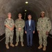Four individuals pose for a formal photograph inside a large concrete tunnel. Three are wearing U.S. Navy NWU Type III (woodland camouflage) uniforms, while one civilian in a blue suit stands among them. The tunnel appears to be a military or government facility with arched concrete walls and overhead lighting receding into the distance.