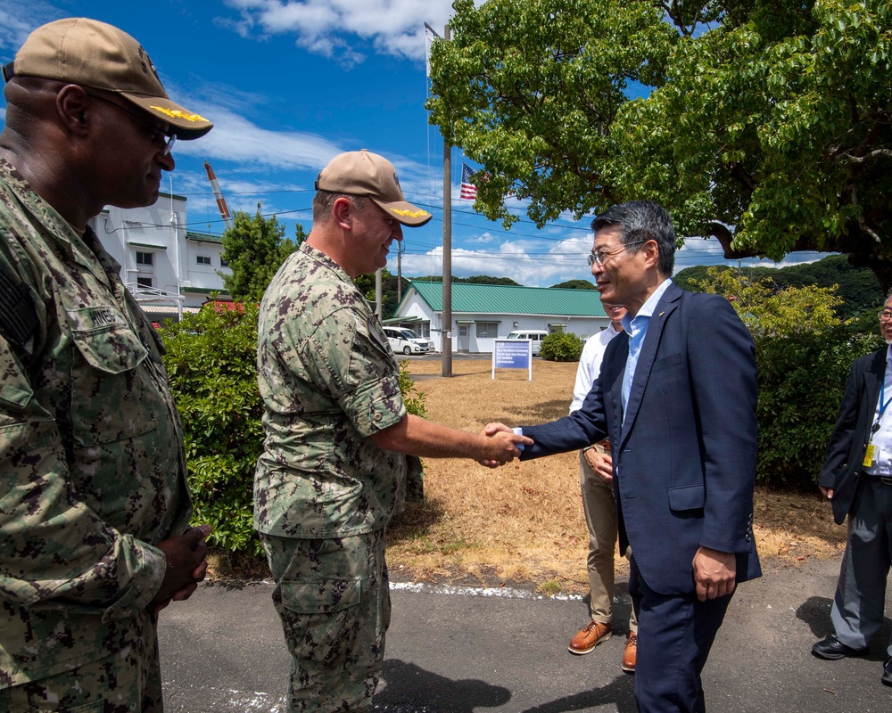 Two U.S. Navy personnel in digital camouflage uniforms are greeting a civilian man in a dark suit outdoors at what appears to be a U.S. military installation. A handshake is taking place between one uniformed service member and the suited civilian, suggesting an official meeting or visit. An American flag is visible in the background, and additional civilians are present on the right side of the frame.