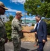 Two U.S. Navy personnel in digital camouflage uniforms are greeting a civilian man in a dark suit outdoors at what appears to be a U.S. military installation. A handshake is taking place between one uniformed service member and the suited civilian, suggesting an official meeting or visit. An American flag is visible in the background, and additional civilians are present on the right side of the frame.