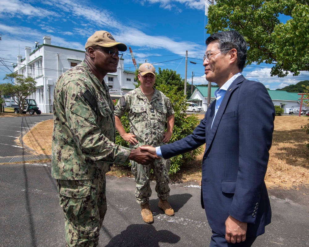 Two U.S. Navy personnel in camouflage uniforms are meeting with a civilian man in a dark business suit outdoors on what appears to be a military installation. The primary interaction is a formal handshake between a senior Navy officer and the civilian, while a second uniformed service member observes in the background. The setting includes military-style white buildings, trees, and a paved area consistent with a U.S. naval base, likely in Japan based on architectural and environmental cues.