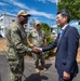 Two U.S. Navy personnel in camouflage uniforms are meeting with a civilian man in a dark business suit outdoors on what appears to be a military installation. The primary interaction is a formal handshake between a senior Navy officer and the civilian, while a second uniformed service member observes in the background. The setting includes military-style white buildings, trees, and a paved area consistent with a U.S. naval base, likely in Japan based on architectural and environmental cues.