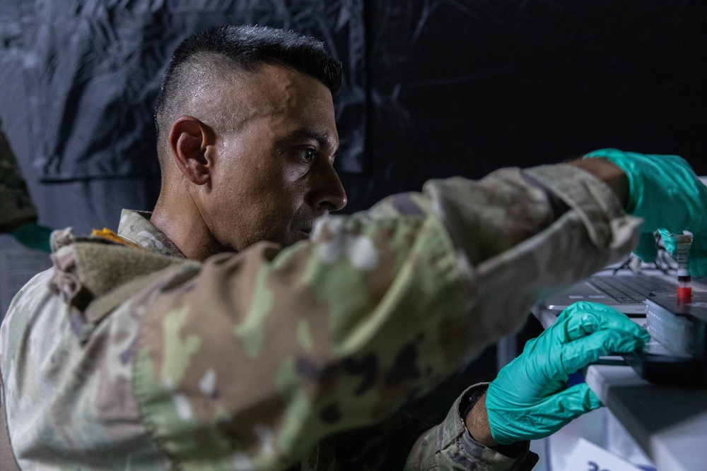 A military personnel member wearing camouflage uniform and green latex gloves is working with what appears to be medical or laboratory specimens/vials on a tray in a dimly lit environment. The individual appears focused and is handling small containers, suggesting a medical, diagnostic, or field laboratory procedure. The setting appears to be a temporary or field medical facility based on the dark backdrop.