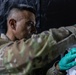 A military personnel member wearing camouflage uniform and green latex gloves is working with what appears to be medical or laboratory specimens/vials on a tray in a dimly lit environment. The individual appears focused and is handling small containers, suggesting a medical, diagnostic, or field laboratory procedure. The setting appears to be a temporary or field medical facility based on the dark backdrop.